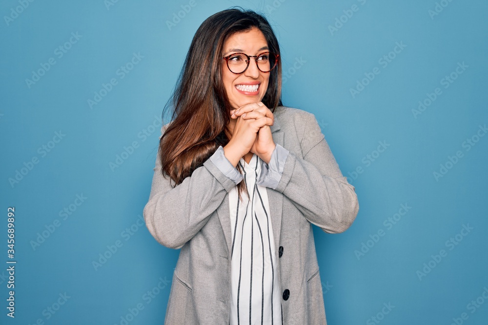 Young hispanic business woman wearing glasses standing over blue isolated background laughing nervous and excited with hands on chin looking to the side Young hispanic business woman wearing glasses standing over blue isolated background laughing nervous and excited with hands on chin looking to the side