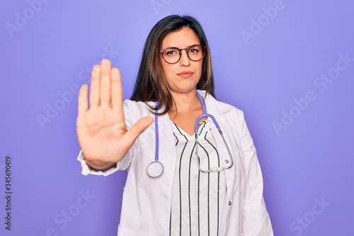 Professional doctor woman wearing stethoscope and medical coat over purple background doing stop sing with palm of the hand. Warning expression with negative and serious gesture on the face.
