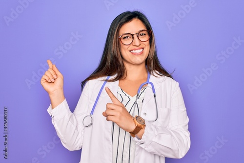 Professional doctor woman wearing stethoscope and medical coat over purple background smiling and looking at the camera pointing with two hands and fingers to the side.