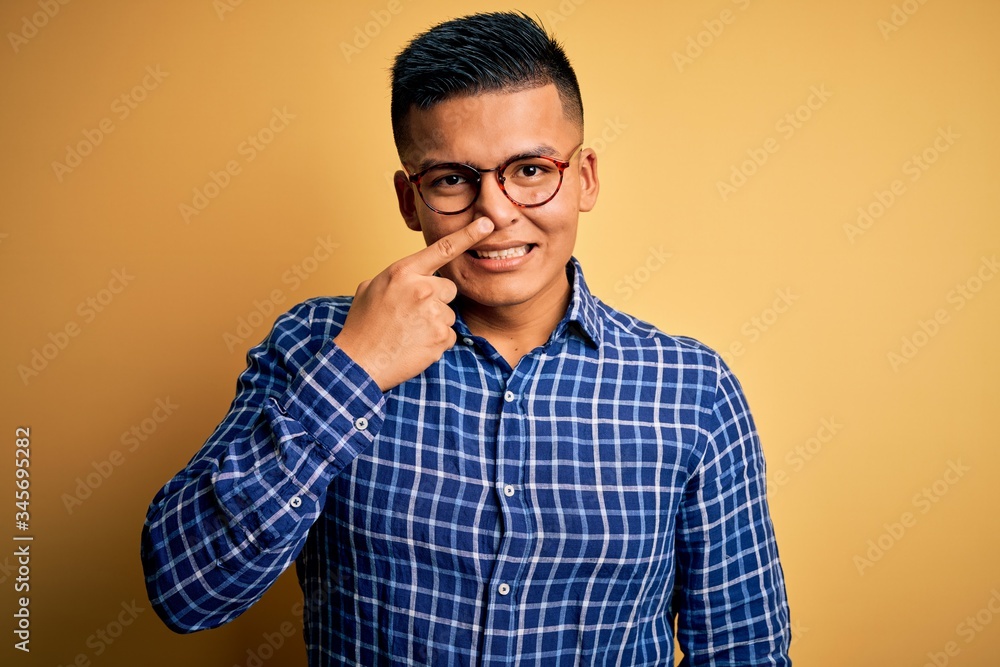 Young handsome latin man wearing casual shirt and glasses over yellow background Pointing with hand finger to face and nose, smiling cheerful. Beauty concept Young handsome latin man wearing casual shirt and glasses over yellow background Pointing with hand finger to face and nose, smiling cheerful. Beauty concept