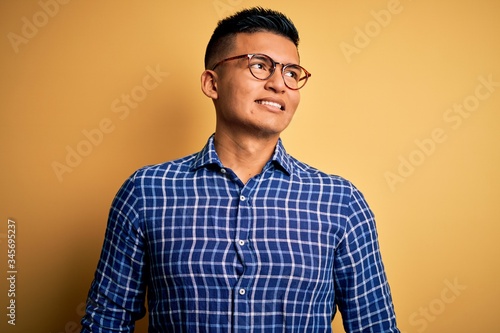Young handsome latin man wearing casual shirt and glasses over yellow background looking away to side with smile on face, natural expression. Laughing confident.
