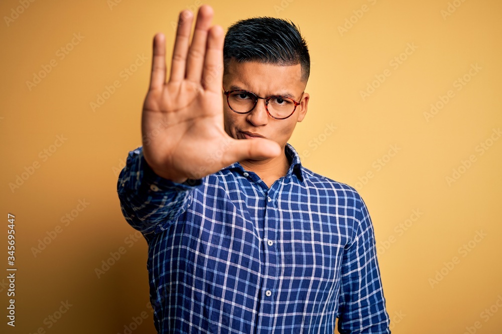 Young handsome latin man wearing casual shirt and glasses over yellow background doing stop sing with palm of the hand. Warning expression with negative and serious gesture on the face. Young handsome latin man wearing casual shirt and glasses over yellow background doing stop sing with palm of the hand. Warning expression with negative and serious gesture on the face.