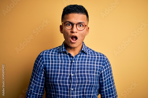 Young handsome latin man wearing casual shirt and glasses over yellow background afraid and shocked with surprise and amazed expression, fear and excited face.