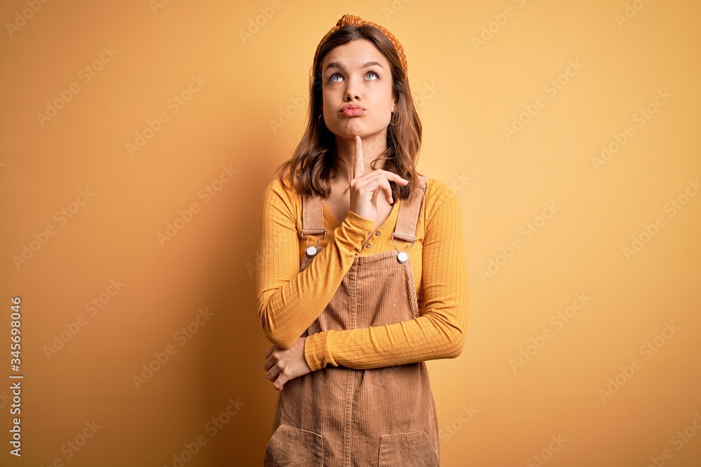 Young beautiful blonde girl wearing overall standing over yellow isolated background Thinking concentrated about doubt with finger on chin and looking up wondering Young beautiful blonde girl wearing overall standing over yellow isolated background Thinking concentrated about doubt with finger on chin and looking up wondering