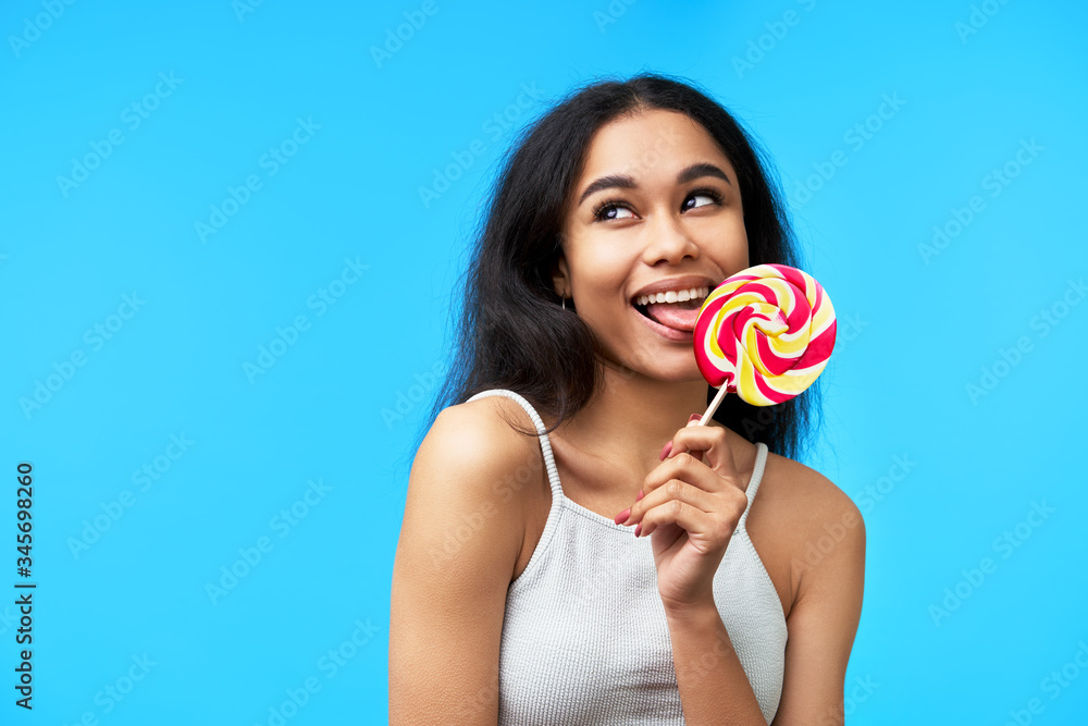 Happy young  black woman licking colorful lollipop on blue background