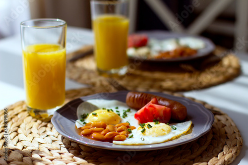 Breakfast on plates of fried eggs, beans, sausages, tomatoes, orange juice in a glass on a table.