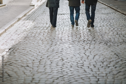 Three men in dark jeans and boots walk along a cobblestone road. The view from the back. The concept of a walk in the old city.