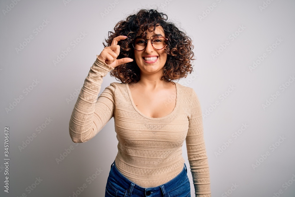 Young beautiful curly arab woman wearing casual t-shirt and glasses over white background smiling and confident gesturing with hand doing small size sign with fingers looking and the camera. Measure Young beautiful curly arab woman wearing casual t-shirt and glasses over white background smiling and confident gesturing with hand doing small size sign with fingers looking and the camera. Measure