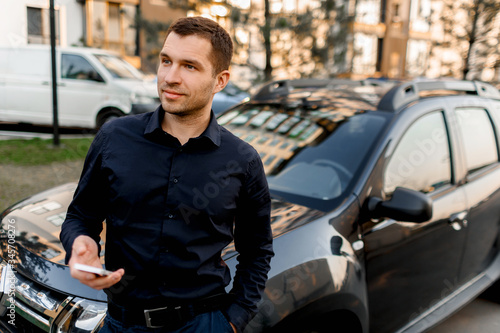 A young man or businessman in a dark shirt stands on the street near the car, looks into the distance in a residential area of the city. The driver is waiting for his passenger or client. © Vlad