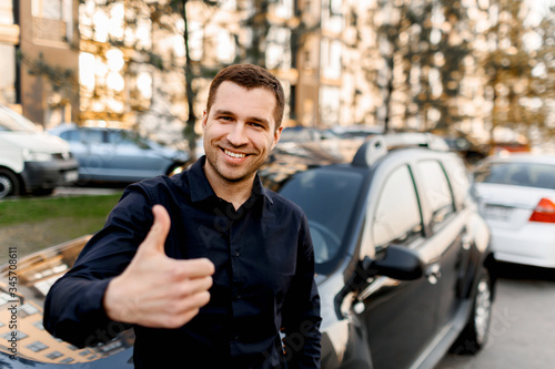 A young man stands near a car and shows a thumb up.Taxi driver looks at the camera and smiles. Urban environment and traffic