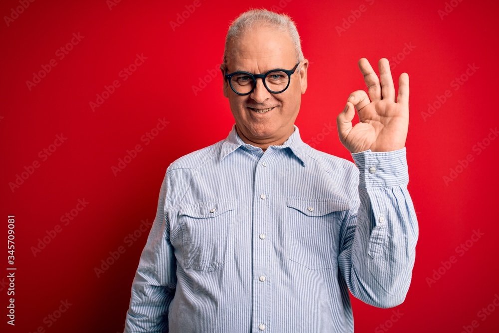 Naklejka premium Middle age handsome hoary man wearing casual striped shirt and glasses over red background smiling positive doing ok sign with hand and fingers. Successful expression.