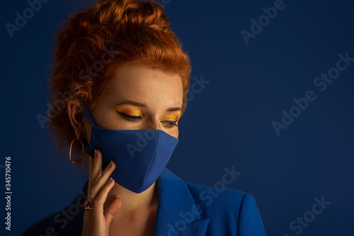 Redhead woman wearing trendy fashion blue monochrome outfit with luxury designer protective face mask. Model has matching bold eyes makeup. Vogue, style during quarantine of coronavirus outbreak. 
