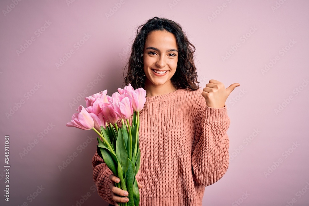 Young beautiful romantic woman with curly hair holding bouquet of pink tulips Pointing to the back behind with hand and thumbs up, smiling confident Young beautiful romantic woman with curly hair holding bouquet of pink tulips Pointing to the back behind with hand and thumbs up, smiling confident