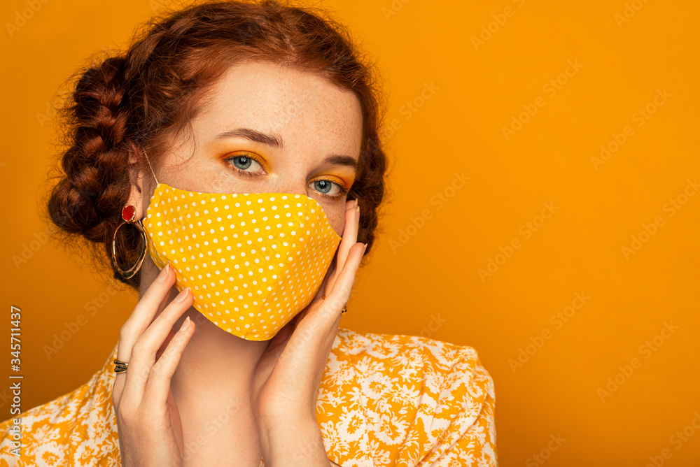 Woman wearing stylish handmade protective face mask posing on orange background.  Model with colorful eyes makeup. Fashion during quarantine of coronavirus outbreak. Copy, empty space for text - obrazy, fototapety, plakaty 