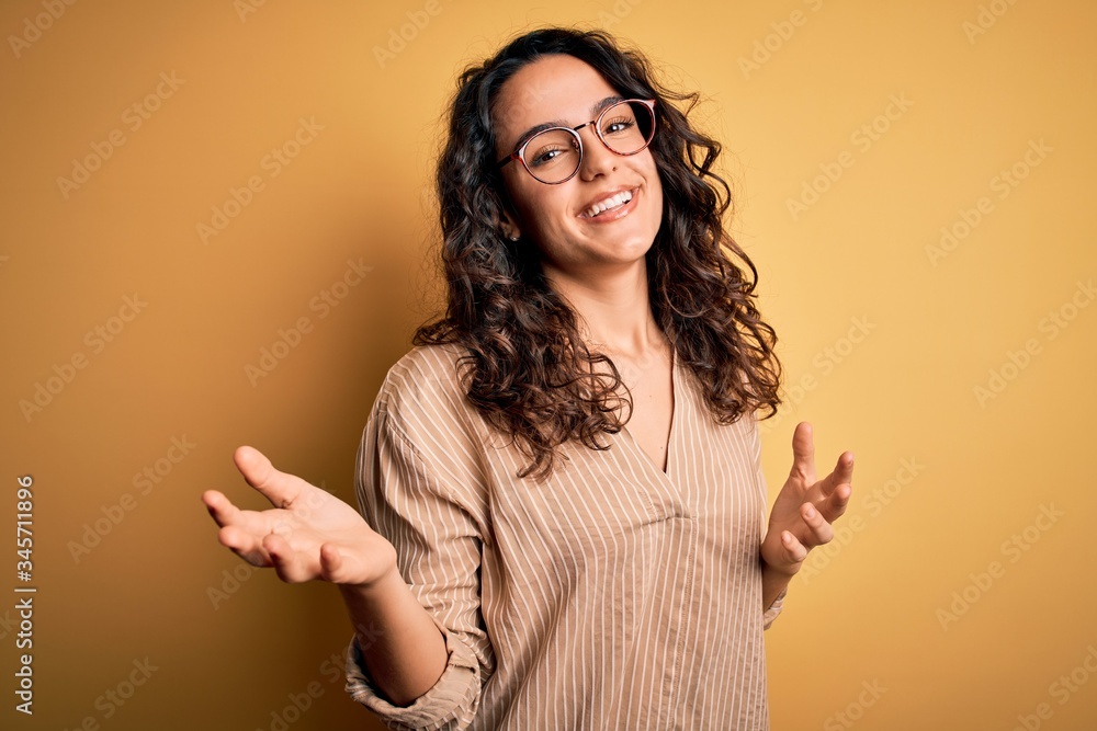 Beautiful woman with curly hair wearing striped shirt and glasses over yellow background smiling cheerful with open arms as friendly welcome, positive and confident greetings Beautiful woman with curly hair wearing striped shirt and glasses over yellow background smiling cheerful with open arms as friendly welcome, positive and confident greetings