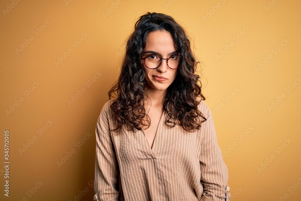 Beautiful woman with curly hair wearing striped shirt and glasses over yellow background skeptic and nervous, frowning upset because of problem. Negative person. Beautiful woman with curly hair wearing striped shirt and glasses over yellow background skeptic and nervous, frowning upset because of problem. Negative person.