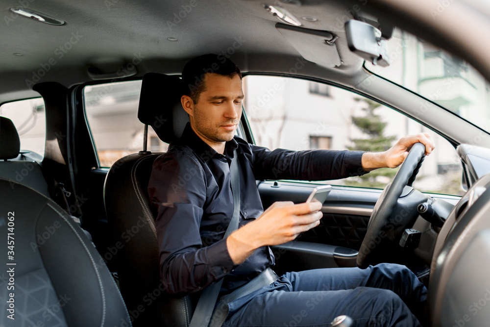 A young man in a dark shirt driving his own car uses a smartphone or phone. Multitasking concept