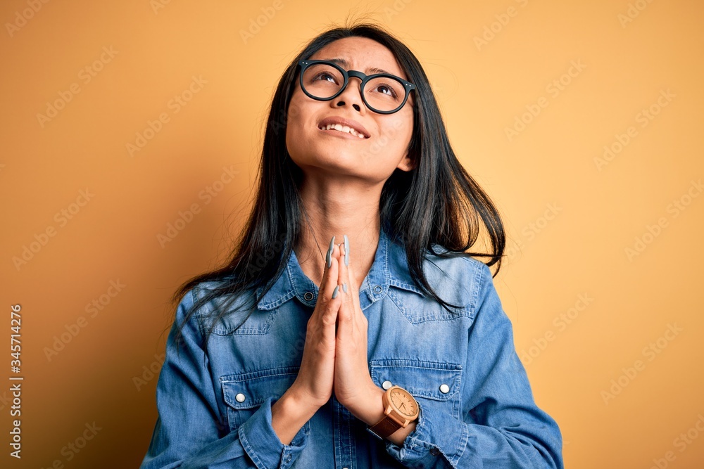 Young beautiful chinese woman wearing casual denim shirt over isolated yellow background begging and praying with hands together with hope expression on face very emotional and worried. Begging.