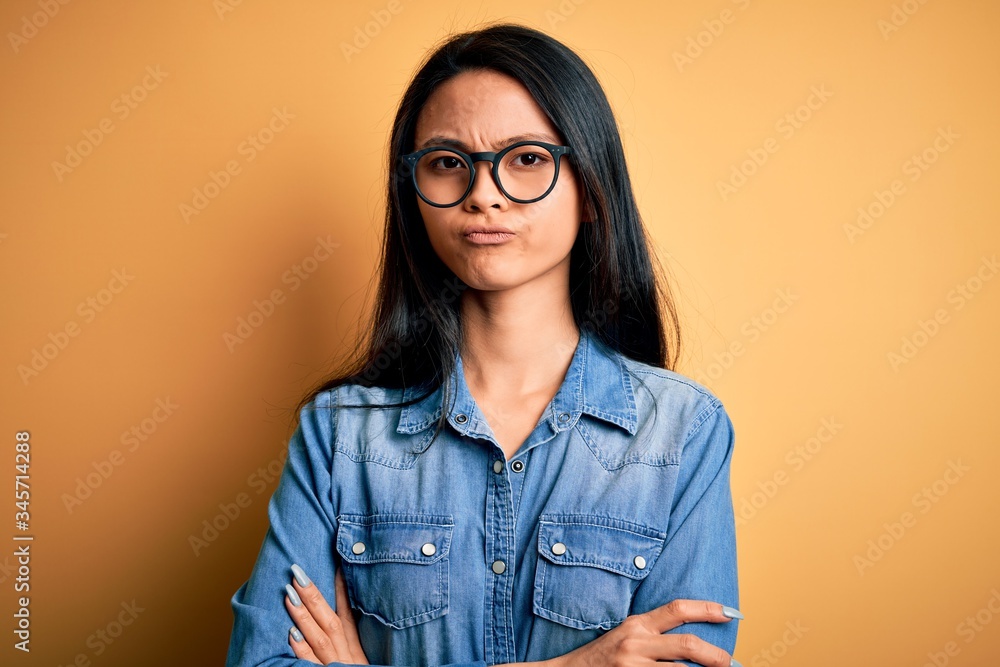Young beautiful chinese woman wearing casual denim shirt over isolated yellow background skeptic and nervous, disapproving expression on face with crossed arms. Negative person.