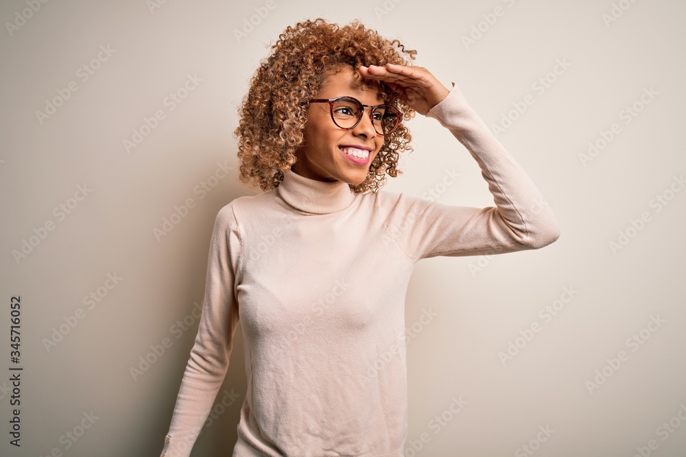Young african american woman wearing turtleneck sweater and glasses over white background very happy and smiling looking far away with hand over head. Searching concept. Young african american woman wearing turtleneck sweater and glasses over white background very happy and smiling looking far away with hand over head. Searching concept.