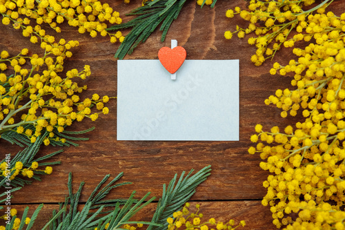 A frame of yellow Mimosa flowers with green leaves with an empty paper card, copy space, flat lay, and a paper clip with a red heart on a brown wooden background