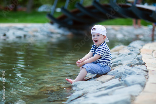Fototapeta Naklejka Na Ścianę i Meble -   child in a white cap in admiration sitting by the pond touches the water with his leg