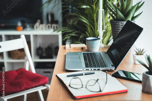 Close up view of a wooden desk with laptop, planner, mobile phone and eyeglasses left on the work table. Work from home concept with window light and green plants during quarantine and isolation