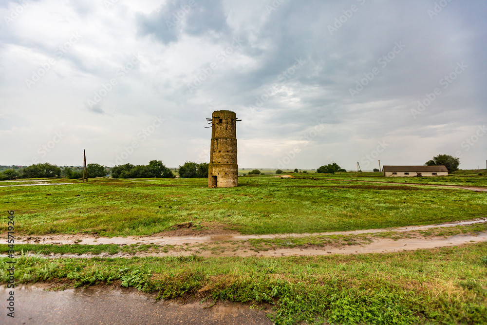 Old brick tower near a sandy road and puddles in a meadow with green grass on a background of cloudy sky. Horizontal orientation.