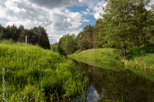 A bend of a small river flowing between green, grassy hills and trees on a warm spring day