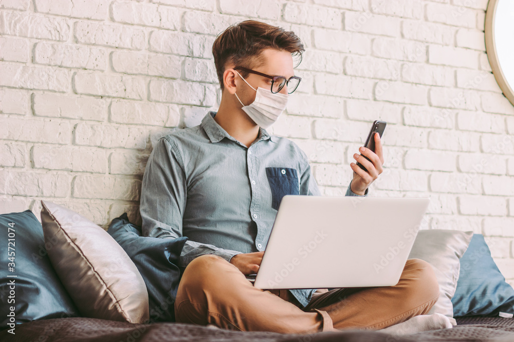 Young businessman in medical face mask using smart phone, texting message while working with laptop at home office. Happy hipster man hold mobile phone in hand, sit on couch. Quarantine communication Young businessman in medical face mask using smart phone, texting message while working with laptop at home office. Happy hipster man hold mobile phone in hand, sit on couch. Quarantine communication