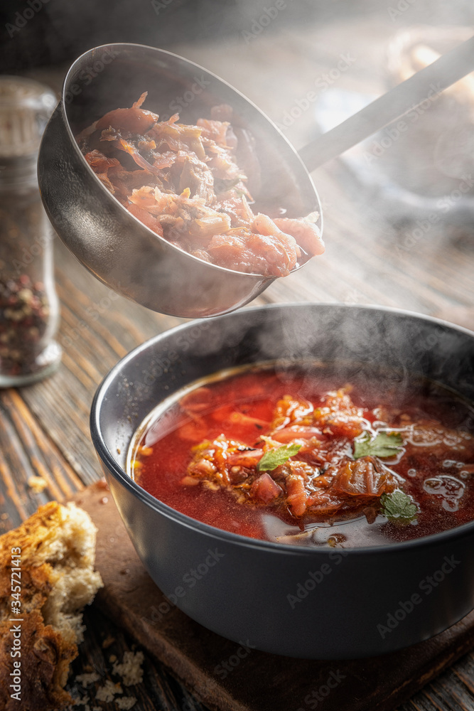 Borsch is poured into a bowl from a ladle from which steam, a traditional Ukrainian vegetable soup made from beets, carrots, tomatoes, potatoes, cabbage, herbs and garlic, close-up, shallow depth of Borsch is poured into a bowl from a ladle from which steam, a traditional Ukrainian vegetable soup made from beets, carrots, tomatoes, potatoes, cabbage, herbs and garlic, close-up, shallow depth of