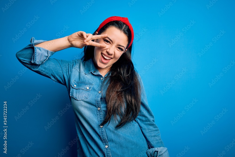 Young brunette woman wearing casual denim shirt over blue isolated background Doing peace symbol with fingers over face, smiling cheerful showing victory Young brunette woman wearing casual denim shirt over blue isolated background Doing peace symbol with fingers over face, smiling cheerful showing victory