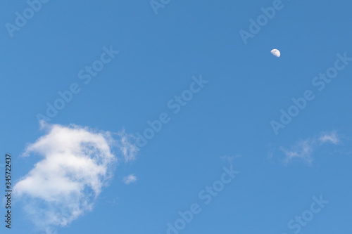 The half moon surround by clouds in the bright blue daytime sky.