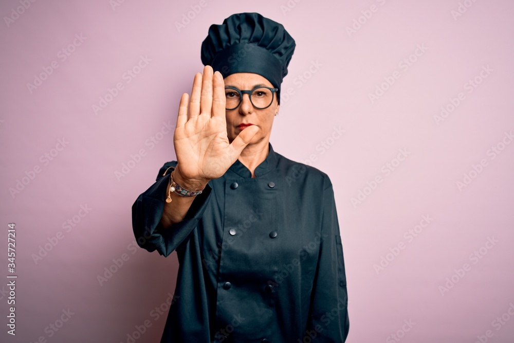 Middle age brunette chef woman wearing cooker uniform and hat over isolated pink background doing stop sing with palm of the hand. Warning expression with negative and serious gesture on the face. Middle age brunette chef woman wearing cooker uniform and hat over isolated pink background doing stop sing with palm of the hand. Warning expression with negative and serious gesture on the face.