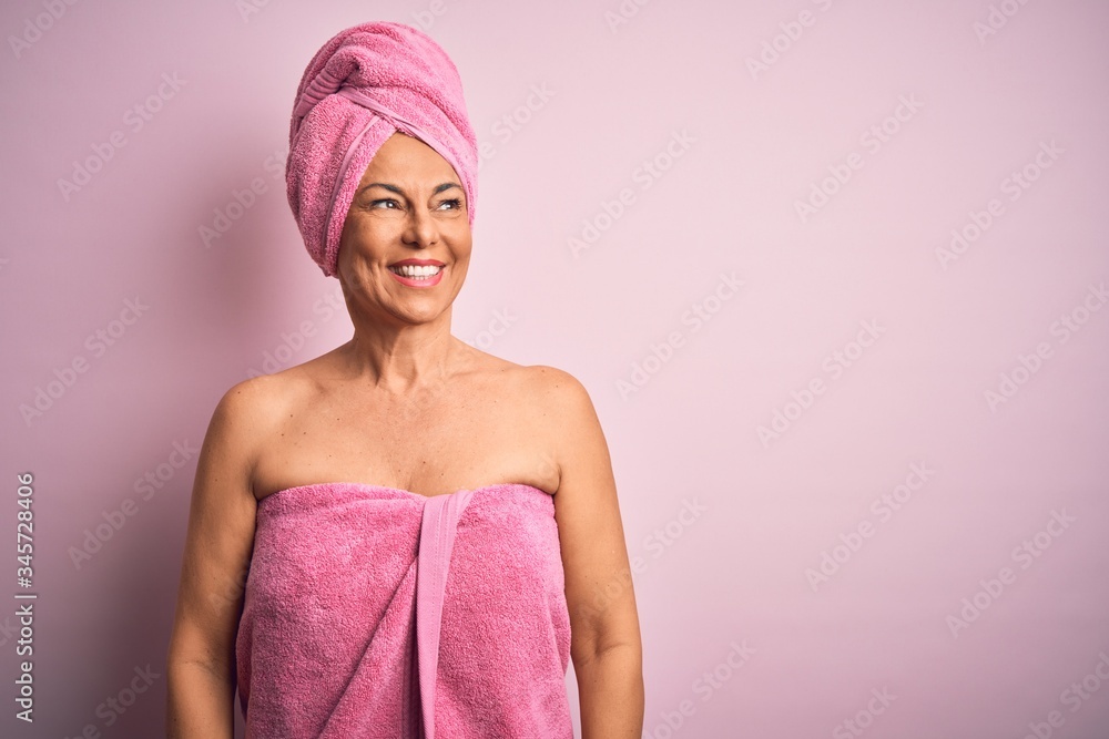 Middle age woman wearing bath towel from beauty body care over pink background looking away to side with smile on face, natural expression. Laughing confident. Middle age woman wearing bath towel from beauty body care over pink background looking away to side with smile on face, natural expression. Laughing confident.