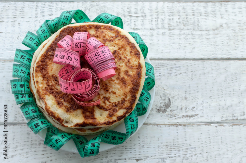 a stack of pancakes on a plate with a centimeter tape on top on a wooden table. the concept of an unhealthy lifestyle. overweight. a stack of pancakes on a plate with a centimeter tape on top on a wooden table. the concept of an unhealthy lifestyle. overweight.