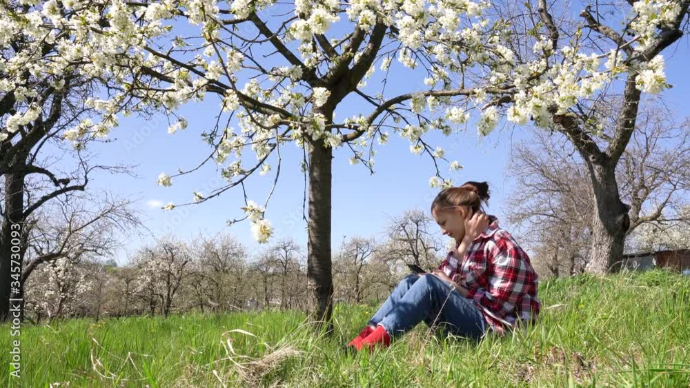 Pretty brunette girl in plaid shirt sitting among blooming garden with phone in her hands Pretty brunette girl in plaid shirt sitting among blooming garden with phone in her hands