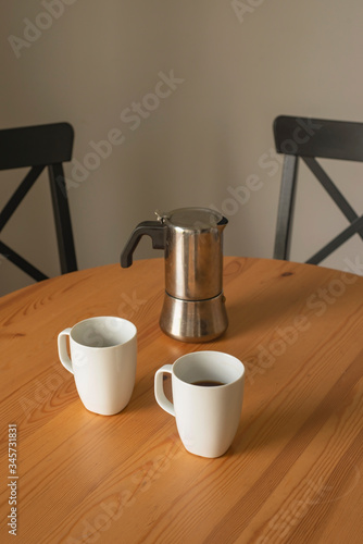 coffee morning at home vertical close-up; two white coffee mugs and a coffee maker over a wooden table with black chairs and a white wall