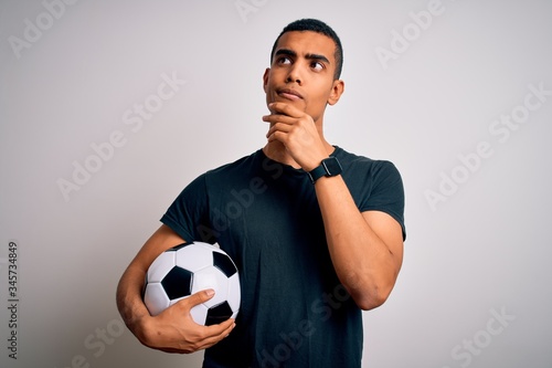 Handsome african american man playing footbal holding soccer ball over white background with hand on chin thinking about question, pensive expression. Smiling with thoughtful face. Doubt concept.