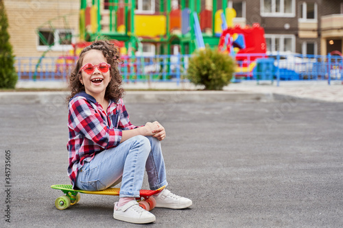 beautiful girl in a plaid shirt on a Playground on a skateboard. a girl with curly hair and pink glasses laughs. Moving activity