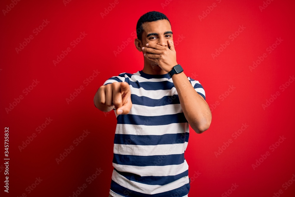 Fototapeta premium Handsome african american man wearing casual striped t-shirt standing over red background laughing at you, pointing finger to the camera with hand over mouth, shame expression
