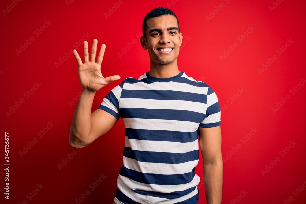 Naklejka premium Handsome african american man wearing casual striped t-shirt standing over red background showing and pointing up with fingers number five while smiling confident and happy.
