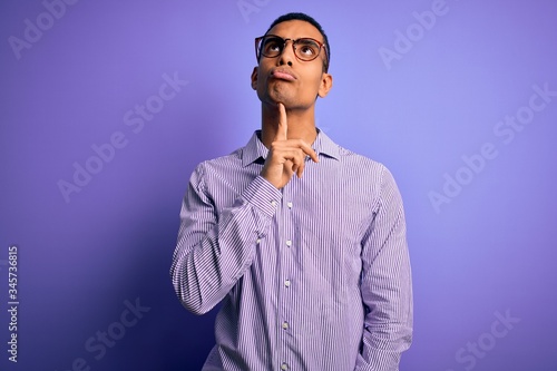 Handsome african american man wearing striped shirt and glasses over purple background Thinking concentrated about doubt with finger on chin and looking up wondering