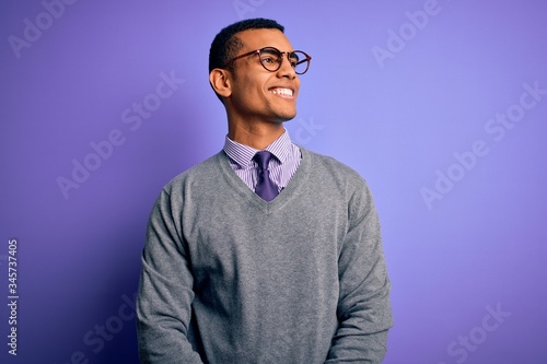 Handsome african american businessman wearing glasses and tie over purple background looking away to side with smile on face, natural expression. Laughing confident.