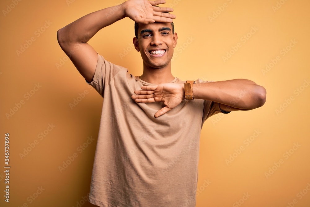 Young handsome african american man wearing casual t-shirt standing over yellow background Smiling cheerful playing peek a boo with hands showing face. Surprised and exited Young handsome african american man wearing casual t-shirt standing over yellow background Smiling cheerful playing peek a boo with hands showing face. Surprised and exited