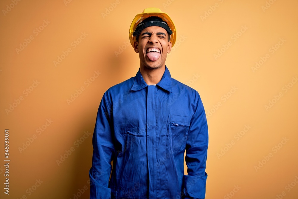 Young handsome african american worker man wearing blue uniform and security helmet sticking tongue out happy with funny expression. Emotion concept. Young handsome african american worker man wearing blue uniform and security helmet sticking tongue out happy with funny expression. Emotion concept.