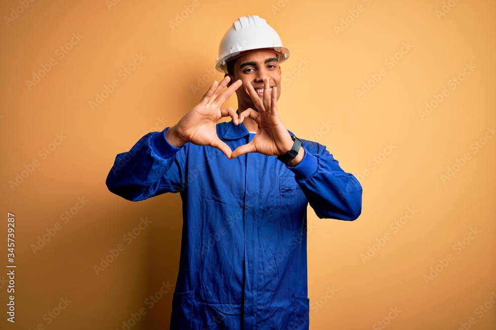 Young handsome african american worker man wearing blue uniform and security helmet smiling in love showing heart symbol and shape with hands. Romantic concept.