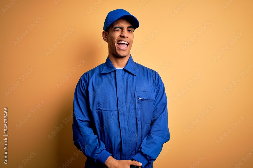 Young african american mechanic man wearing blue uniform and cap over yellow background winking looking at the camera with sexy expression, cheerful and happy face.