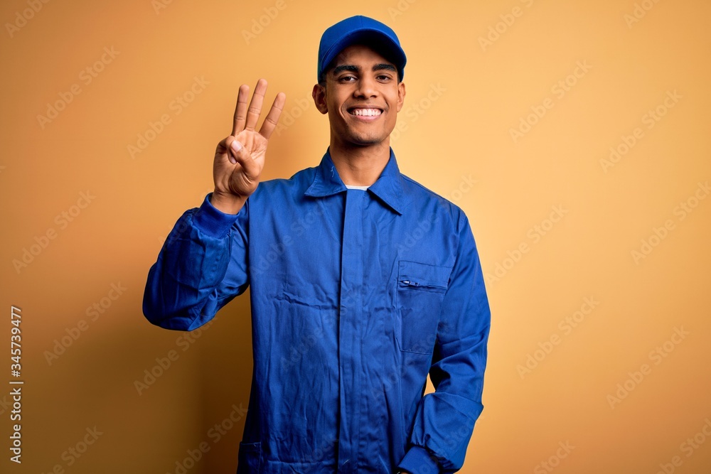 Young african american mechanic man wearing blue uniform and cap over yellow background showing and pointing up with fingers number three while smiling confident and happy.
