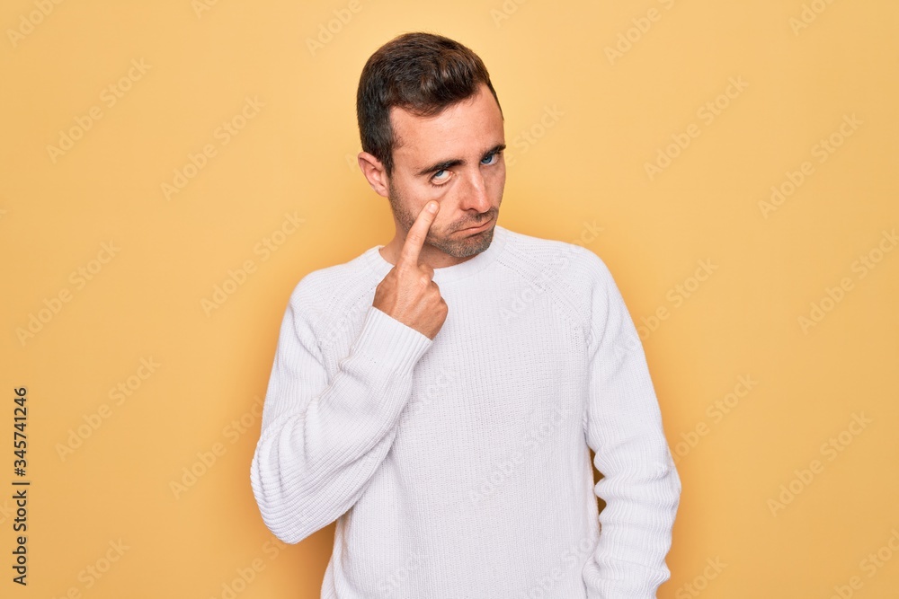 Young handsome man with blue eyes wearing casual sweater standing over yellow background Pointing to the eye watching you gesture, suspicious expression Young handsome man with blue eyes wearing casual sweater standing over yellow background Pointing to the eye watching you gesture, suspicious expression